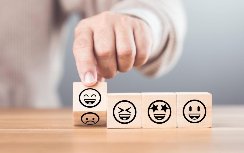 A hand positions a wooden block displaying a smiling emoji among others with various expressions, highlighting emotions in a user feedback context.