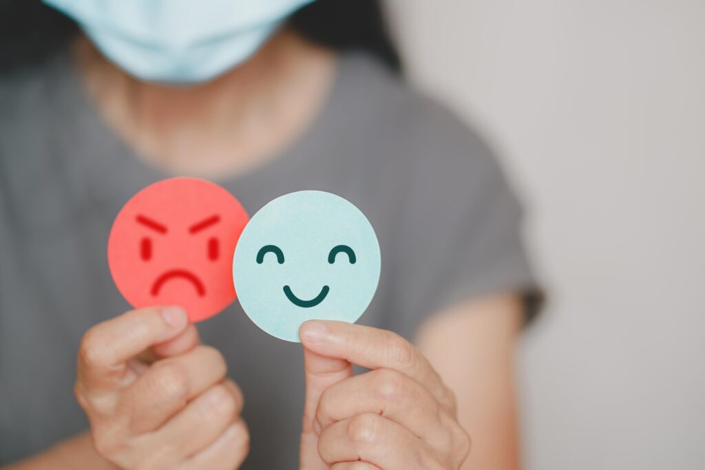A person wearing a mask holds two expression stickers: a red angry face and a blue happy face, illustrating contrasting emotions.
